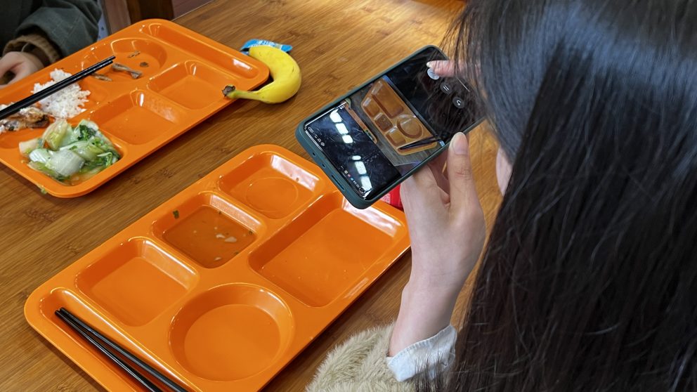 Imagem de uma aluna fotografando uma bandeja laranja de alimentos