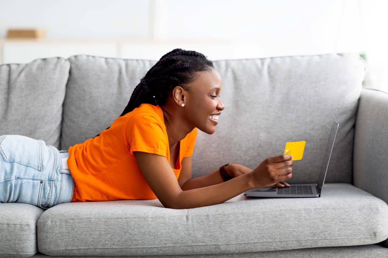 Garota deitada de bruços em um sofá, sorrindo, segurando um cartão de crédito e olhando para um notebook